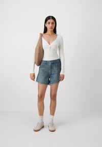Hvit langermet topp med knyttet front, blå denimshorts med frynset kant, beige veske og hvite joggesko med brune såler.