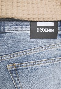 Jean en denim bleu clair avec un tissu texturé couleur beige au-dessus. Présente une étiquette noire avec le logo "DRDENIM" cousu sur la ceinture arrière.
