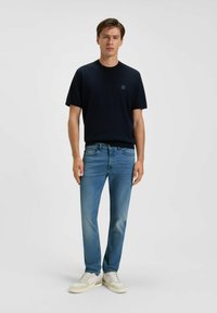 T-shirt di cotone blu navy con maniche corte e un piccolo logo, abbinata a jeans di denim azzurri e sneakers bianche con texture. Design semplice e lineare.