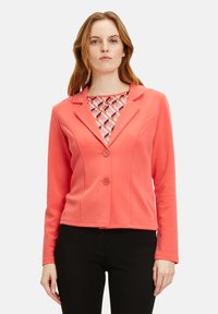 Koralfarbenes Blazer mit langen Ärmeln und Spitzkragen, ausgestattet mit zwei Knöpfen und einer strukturierten Passform. Das Innenshirt hat ein geometrisches Muster.