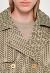 Gehaakte trenchcoat in olijfgroen met een crème visgraatpatroon. Voorzien van grote revers, dubbele knopen en een getextureerde trui eronder.