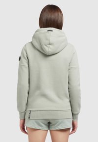 Persona con capelli castani che indossa una felpa con cappuccio grigia chiara e pantaloni corti abbinati, in piedi con la schiena rivolta contro uno sfondo neutro.