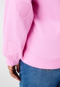 Roze sweatshirt mouw met twee geborduurde hart patches boven een hand die naast blauwe denim jeans rust.