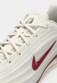 Šviesiai pilki sportiniai bateliai su tekstūruota tinklinė viršutine dalimi, raudonu „swoosh“ logotipu ir smėlio spalvos raišteliais. Yra paminkštintas padas ir kvėpuojanti konstrukcija.