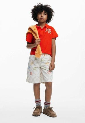 Piros galléros ing hímzett felirattal, bézs rövidnadrág színes virágmintákkal, szürke zoknival és barna cipővel párosítva. Narancssárga pulóvert dobott rá.