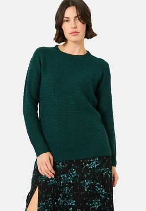 Green knit sweater featuring a textured pattern, round neck, and ribbed cuffs. Worn over a black skirt with a floral print.