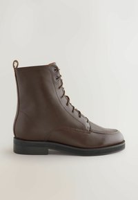 Botine din piele maro până la gleznă cu un design cu șireturi, vârf rotund, cusături contrastante și o talpă din cauciuc negru. Dispune de o mică bucată de plastic pentru tragere în spate.