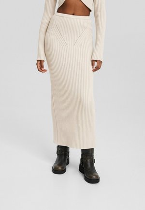 Vrouw draagt een crèmekleurige geribbelde gebreide midi-rok met hoge taille en zwarte gespschoenen op een effen achtergrond.