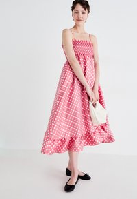Pink polka dot dress with a smocked bodice, thin straps, and a ruffled hem, paired with a white textured bag and black flats.