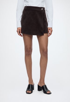 Femme portant une jupe portefeuille en velours côtelé marron foncé, une chemise blanche à manches longues et des mules ouvertes à talons noirs, debout devant un fond uni.
