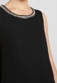 Haut noir sans manches avec un col rond orné d'une bordure perlée argentée et noire, texture lisse et coupe ample.