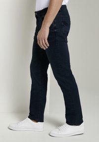 Tmavě modré džíny s rovnými nohavicemi, s předními a zadními kapsami, v kombinaci s bílými teniskami, zobrazené z bočního úhlu.