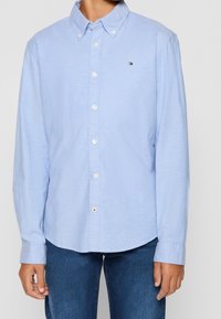 Junge trägt ein hellblaues Hemd mit Knöpfen und Logo auf der Brust sowie dunkle blaue Jeans, steht mit entspannten Armen an den Seiten.