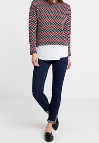 Pull à manches longues à motifs rouge, marine et blanc, porté sur une chemise blanche. Associé à un jean bleu foncé et des ballerines noires.
