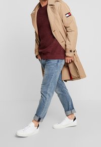 Beige trenssitakki viininpunaisen neuleen päällä, yhdistetty vaaleansinisiin käännettyihin farkkuihin ja valkoisiin lenkkareihin, joissa on mustat yksityiskohdat.