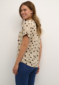 Haut beige à manches courtes avec des pois noirs, coupe décontractée, longueur hanches, tissu léger, ourlet arrondi et texture légèrement brillante.
