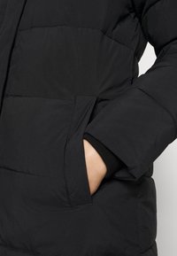 Černá prošívaná bunda s hladkou texturou, která má boční kapsu a žebrované manžety. Design klade důraz na teplo a funkčnost.