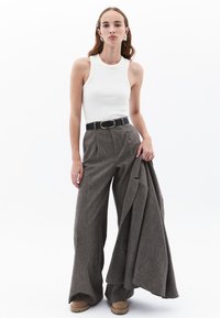 Camisetta bianca aderente, pantaloni a quadri grigi con vita alta, cintura nera con fibbia dorata, mentre tiene un giubbotto grigio, scarpe color cammello. Sfondo neutro.