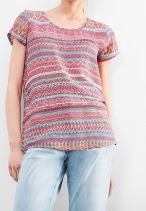 Kvinde iført en kortærmet, mønstret rød og blå top med geometriske designs, kombineret med lyseblå jeans mod en hvid baggrund.