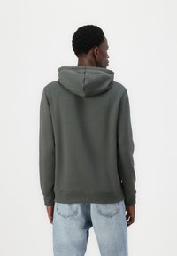 Sweat à capuche vert foncé avec une texture douce et une poche kangourou, présentant des poignets et un ourlet côtelés, porté sur un jean bleu clair.