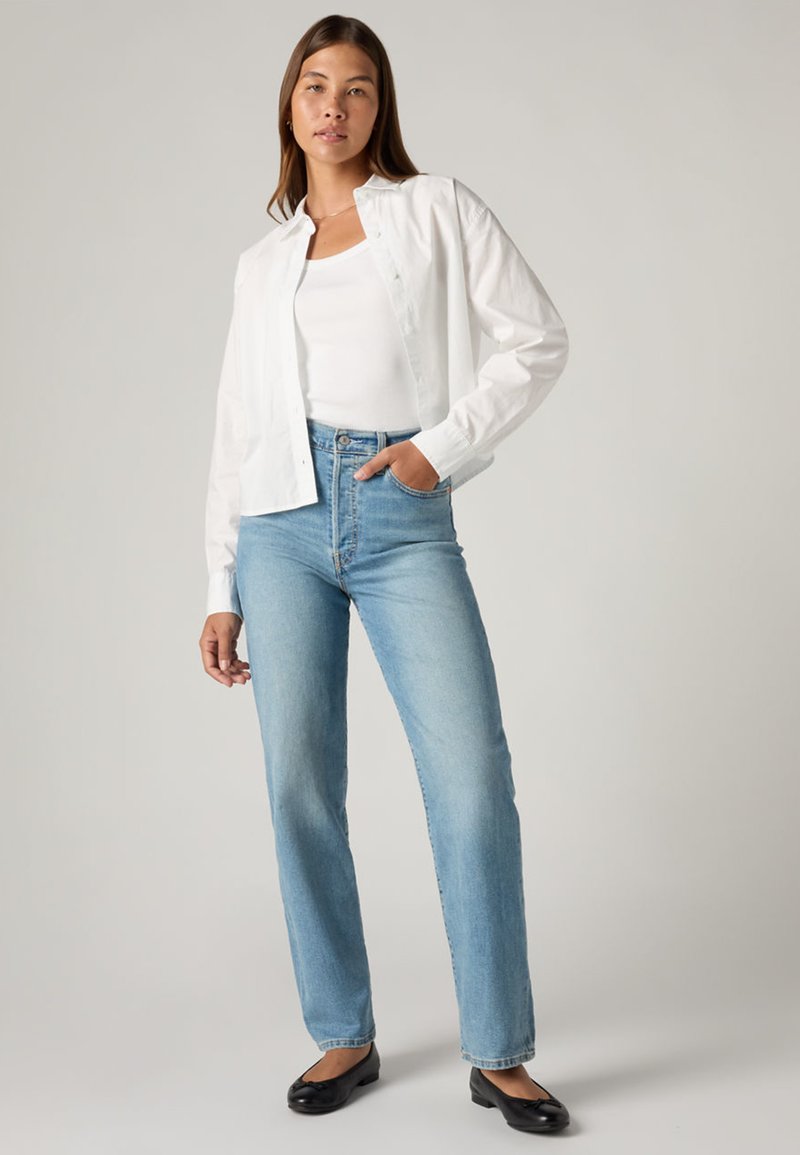 Camicia bianca con bottoni sopra una canottiera bianca, abbinata a jeans dritti azzurri e ballerine nere, su sfondo neutro.