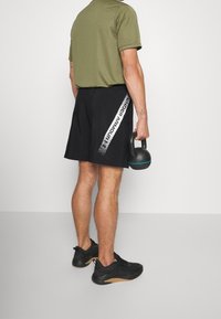 Pantalones cortos deportivos negros con una franja diagonal blanca que presenta el texto "Under Armour"; combinados con una camiseta verde oliva y zapatillas negras.