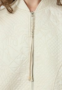 Giacca bianca trapuntata con colletto alto, motivo tessurizzato intricato e chiusura con zip dotata di dettagli decorativi sul tirante.