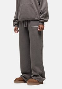 Grijze oversized hoodie met een voorzak, gecombineerd met grijze wijde sweatpants met een wit logo. Model draagt bruine instap schoenen.