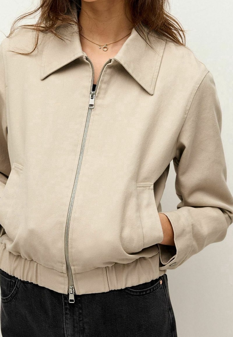Veste zippée beige avec un large col, des poches latérales et une taille élastique. Elle présente un tissu texturé et une fermeture éclair en argent.