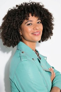 Eine Person mit lockigem Haar lächelt, trägt eine mintgrüne Lederjacke mit silbernem Zubehör über einem Leopardenmuster-Top vor einem weißen Hintergrund.