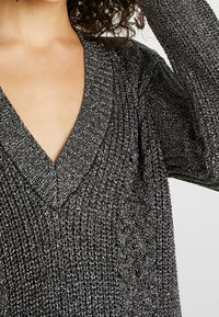 Pull noir étincelant en maille avec un décolleté en V profond, un tissu texturé et des manches longues. Accents de brillance sur toute la surface.