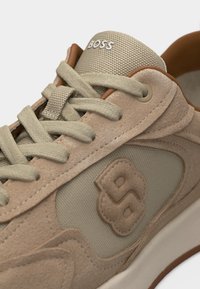 Κοντινό πλάνο αθλητικού παπουτσιού από μπεζ σουέντ και δίχτυ με μπεζ κορδόνια, λογότυπο «BOSS» στη γλώσσα και ραμμένο έμβλημα «B» στο πλάι.