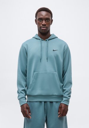 Vyras, vilkintis tamsiai žalią Nike džemperį su kapišonu ir derančius sportinius kelnes, stovintis prie paprasto šviesaus fono, veidas nukreiptas tiesiai į priekį, neutralus išraiškas.