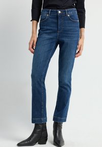 Vaqueros de denim azul oscuro con una cintura alta, corte recto y sutiles detalles de costura. Combinados con botas de tobillo negras.
