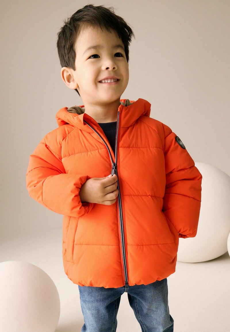 Jeune garçon souriant en train de zipper une doudoune orange, portant un jean bleu, avec des objets blancs ronds en arrière-plan.