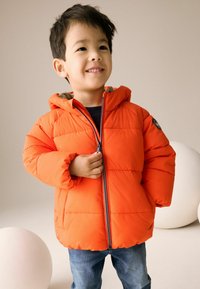 Helles oranges gepolstertes Jacket mit einer Kapuze, ausgestattet mit einem Reißverschluss vorne und Seitentaschen, getragen über einem marineblauen Oberteil und blauen Jeans.