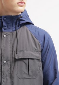 Mand iført en blå og grå hættetrøje med knapper foran og en stor klaplomme på brystet, vist fra nakken til midten af torsoen.