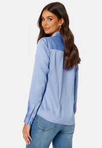 Camicia in satin azzurro chiaro con maniche lunghe, polsino con bottoni e orlo leggermente curvo, con un pannello posteriore di colore più scuro in contrasto.