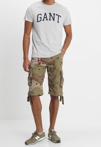 Camiseta gris de manga corta con "GANT" en azul marino, combinada con pantalones cortos cargo de camuflaje que cuentan con múltiples bolsillos y zapatillas deportivas grises.