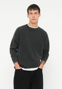 Dunkelgrauer Baumwollpullover mit langen Ärmeln und Rundhalsausschnitt, mit einer weichen Textur und einer lässigen Passform. Über einem weißen T-Shirt getragen.