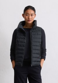 Jeune femme aux cheveux bruns en chignon, portant un gilet sans manches noir matelassé sur un haut noir à manches longues, se tenant devant un mur blanc.