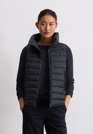Jeune femme aux cheveux bruns en chignon, portant un gilet sans manches noir matelassé sur un haut noir à manches longues, se tenant devant un mur blanc.