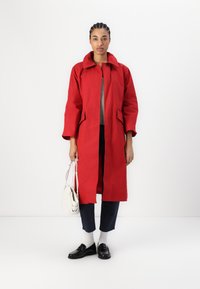 Czerwony wodoodporny płaszcz z kołnierzem, zapinany na przód na guziki, z dwoma bocznymi kieszeniami i prostym krojem. Połączony z ciemnymi spodniami i czarnymi mokasynami.