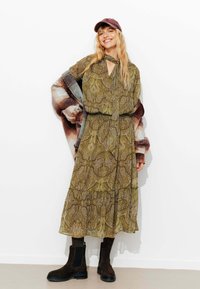 Robe longue verte à motif paisley avec une ceinture à la taille, des volants superposés et un design en trou de serrure. Assortie à un châle à carreaux marron et des bottines noires.