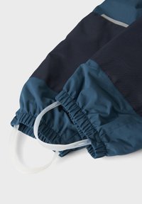 Pantalones impermeables en azul y marino con puños elásticos y estribos ajustables blancos sobre un fondo blanco.