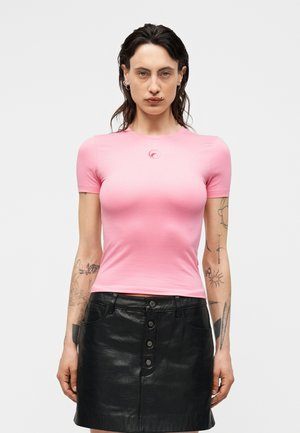 Femeie cu păr întunecat și umed, purtând un tricou roz mulat cu mâneci scurte și o fustă neagră din piele cu nasturi în față, arătând tatuaje pe ambele brațe.