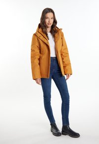 Giacca isolante gialla con cappuccio, apertura con zip e tasche laterali, abbinata a jeans skinny blu scuro e stivaletti neri.