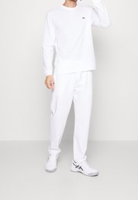 Vit långärmad skjorta, vita sweatpants och ljusgrå sneakers med svarta accenter. Skjortan har en liten grön logotyp på bröstet.