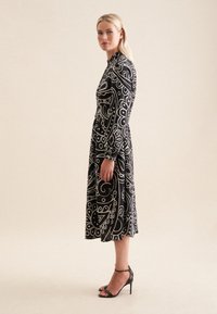 Langes schwarzes Kleid mit weißen wirbelnden Mustern, langen Ärmeln und hohem Kragen. Kombiniert mit schwarzen Riemchensandalen mit Absatz.