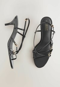 Sorte patentlæder hælede sandaler med tynde stropper, guldfarvet hardware, åben tå-design og en slank, minimalistisk silhuet.